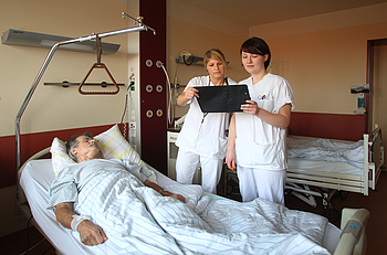 Zwei Krankenschwestern mit Patientenakte am Bett von einem männlichen Patienten.